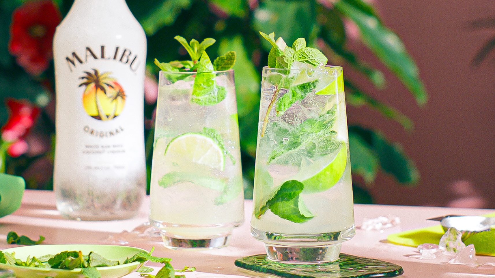 Two Malibu Mojitos on a table in a tropical setting with a bottle of Malibu rum in the background